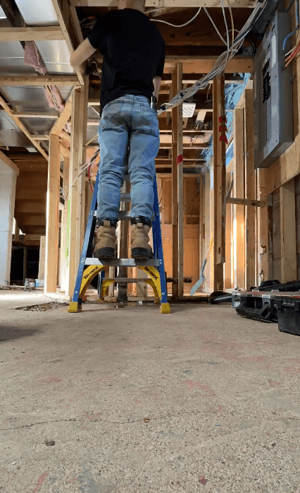 Electrician on a blue ladder installs wiring in an unfinished basement with exposed studs.